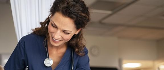 Nurse woman with brunette hair with stethoscope around neck looks down smiling at unseen patient.
