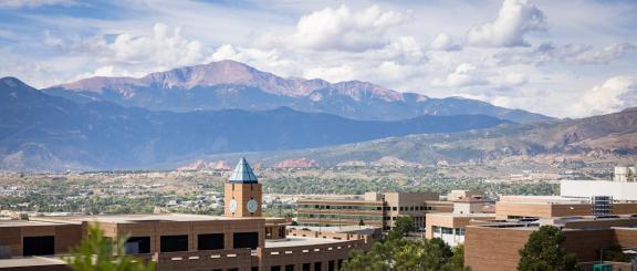 Beautiful blue mountain range with white clouds in the sky. Garden of the Gods red/orange rock formations are seen in the distance, behind the iconic, pointy blue clocktower roof of the University of Colorado campus buildings in the foreground.
