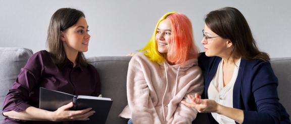 Social worker woman sitting on couch, looking over at concerned mother and smiling daughter (with brightly colored hair) next to her.