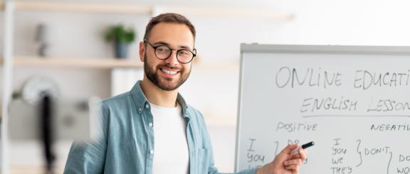 Man smiles at phone camera on tripod in front of him. He points to a dry erase board with "Online Education English Lesson" writing.