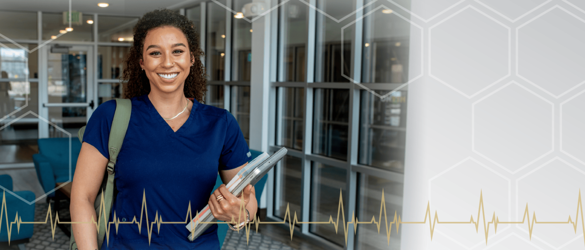 Young woman smiling, wearing nursing scrubs, holding her laptop and a textbook as she walks out of a building