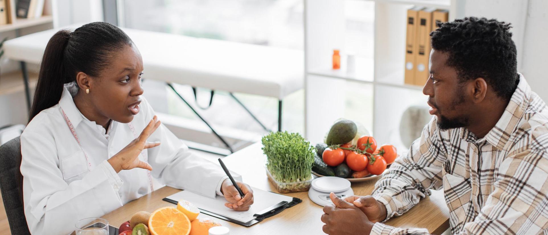 Woman in white with measuring tape hanging around her neck, writing on clipboard, explaining nutrition to man sitting across from her, with vegetables on their table.