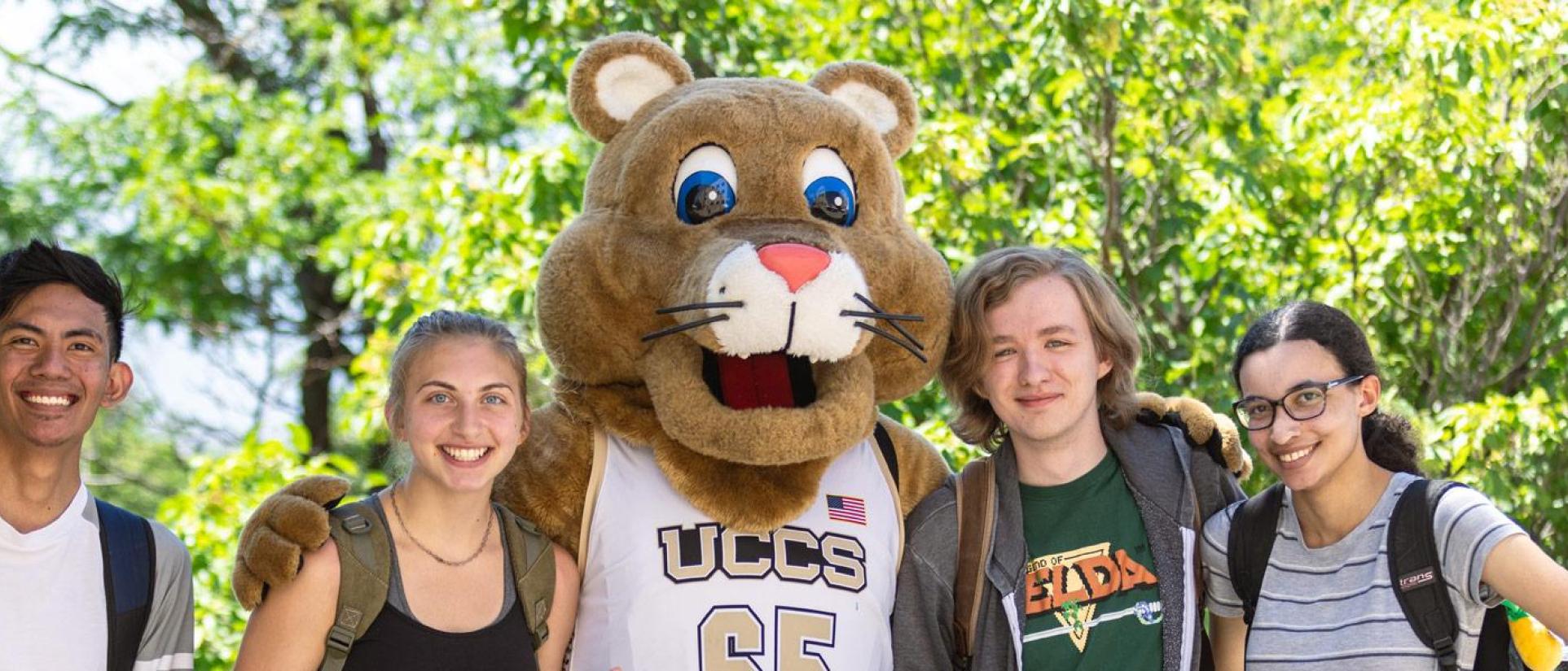 UCCS mascot, Clyde the Mountain Lion, wearing a white and gold basketball jersey, poses with arms around 4 young college students, smiling and wearing backpacks. Behind are bright green trees on a sunny day.