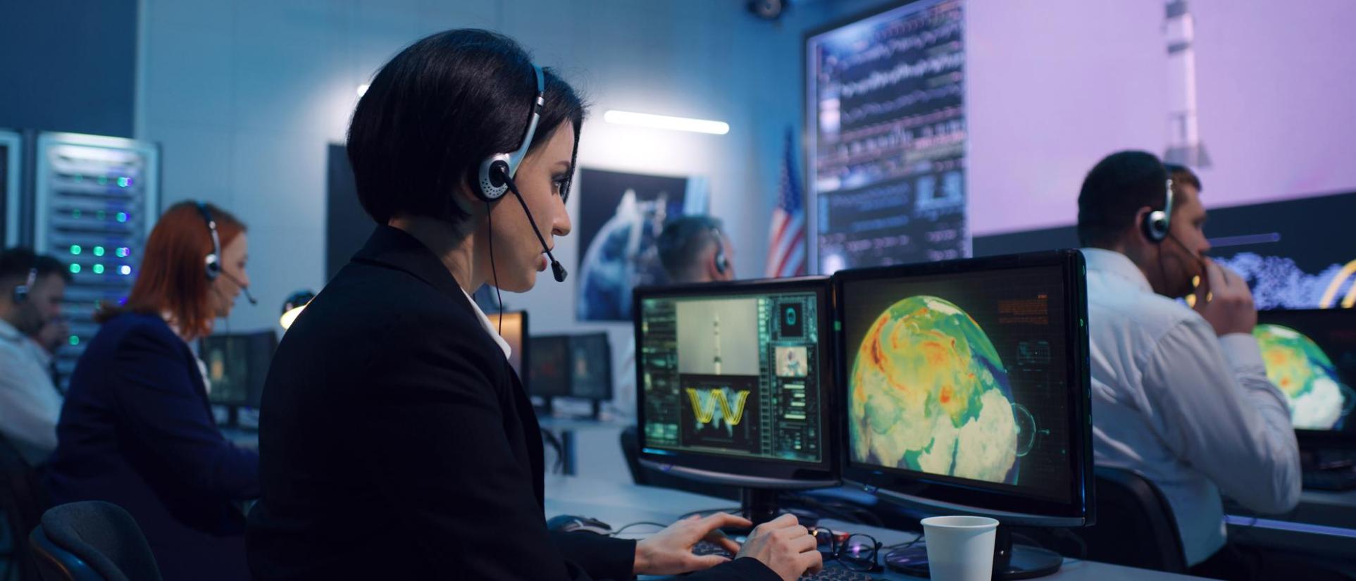 Woman in control room wearing a headset, looking into computer monitor showing planet earth with a large screen in the background showing a rocket being launched.