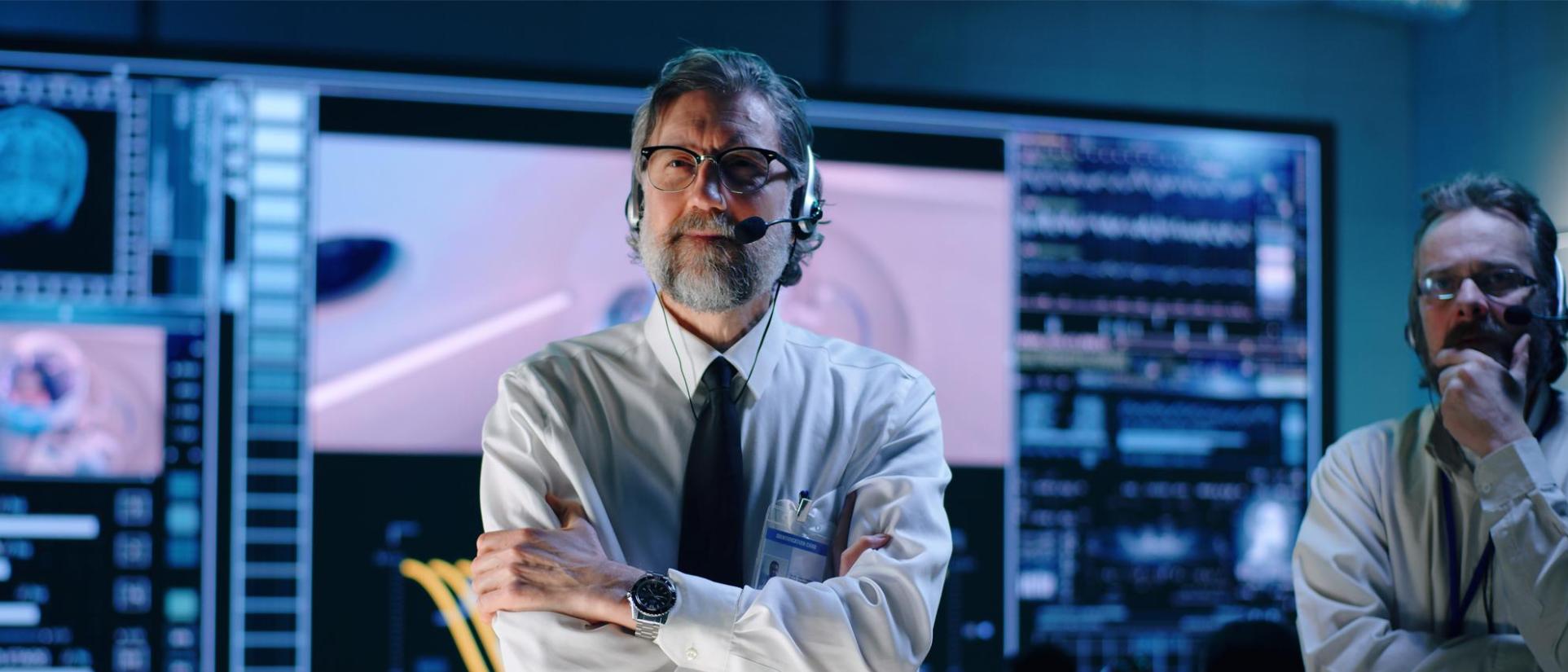 Man with greying beard and glasses, wearing headphones with an attached microphone, ID badge, crisp white shirt, tie and nice watch. His arms are crossed and he looks concerned with a control panel with many large screens behind him. Next to him is a man with his hand on his chin, with a furrowed brow.