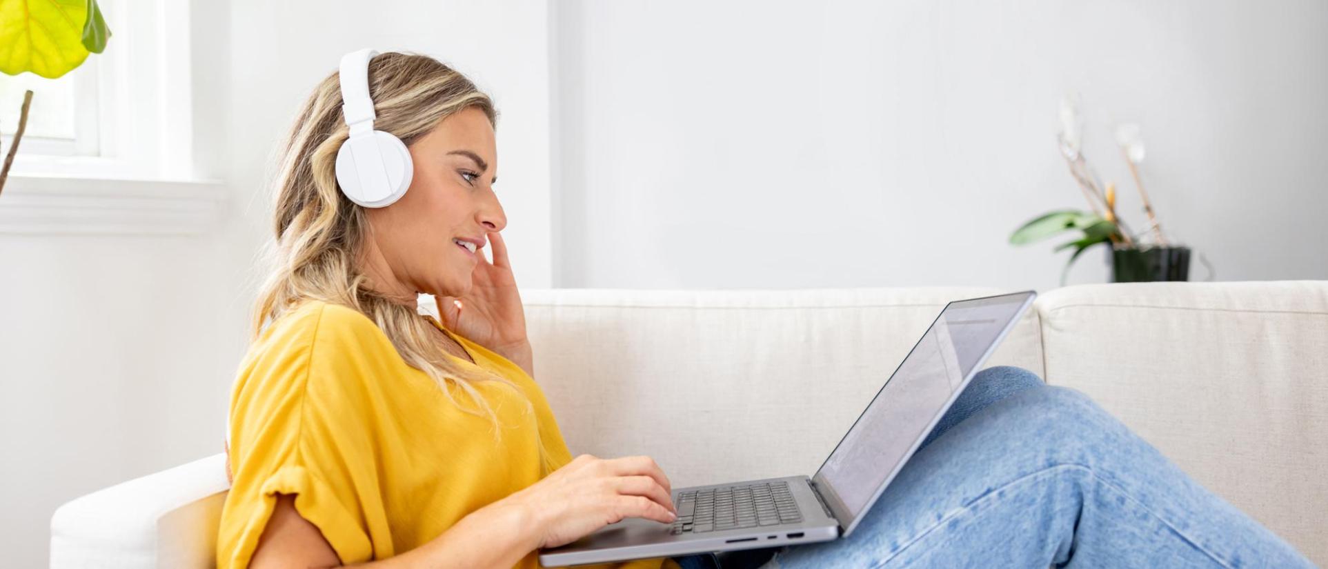 Smiling blonde woman wearing white headphones and gold/yellow shirt, sits on her couch with a laptop in her lap, in a bright home.