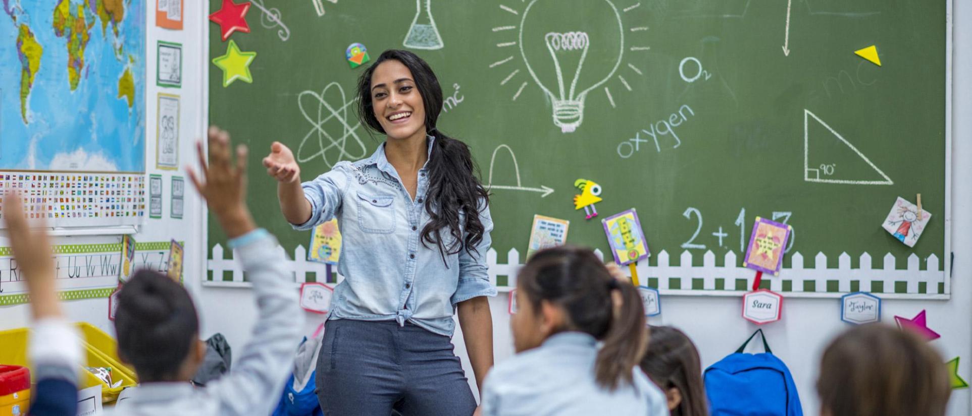 Smiling teacher woman in denim shirt with brunette hair gestures her arm towards an elementary school student boy, raising his hand to be called on. Behind her is a green chalkboard with many symbols drawn in white - such as a light bulb, atom, triangle, graph, science beaker, math equation, ect. and a world map on the wall. Many other young student kids sit in their desks in front of their teacher.