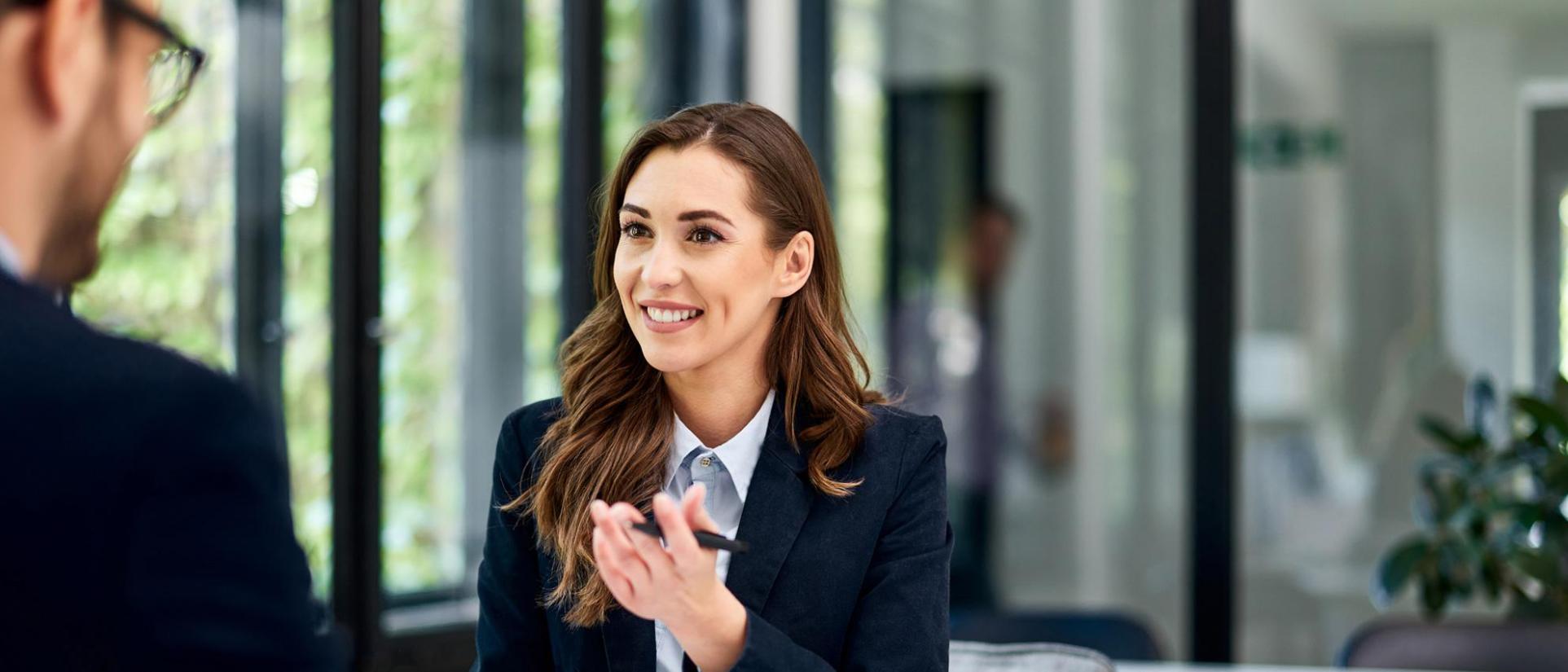 Brunette, long-haired woman wearing professional attire, smiles at man sitting across from her, as she motions her hand (holding a pen) towards him.
