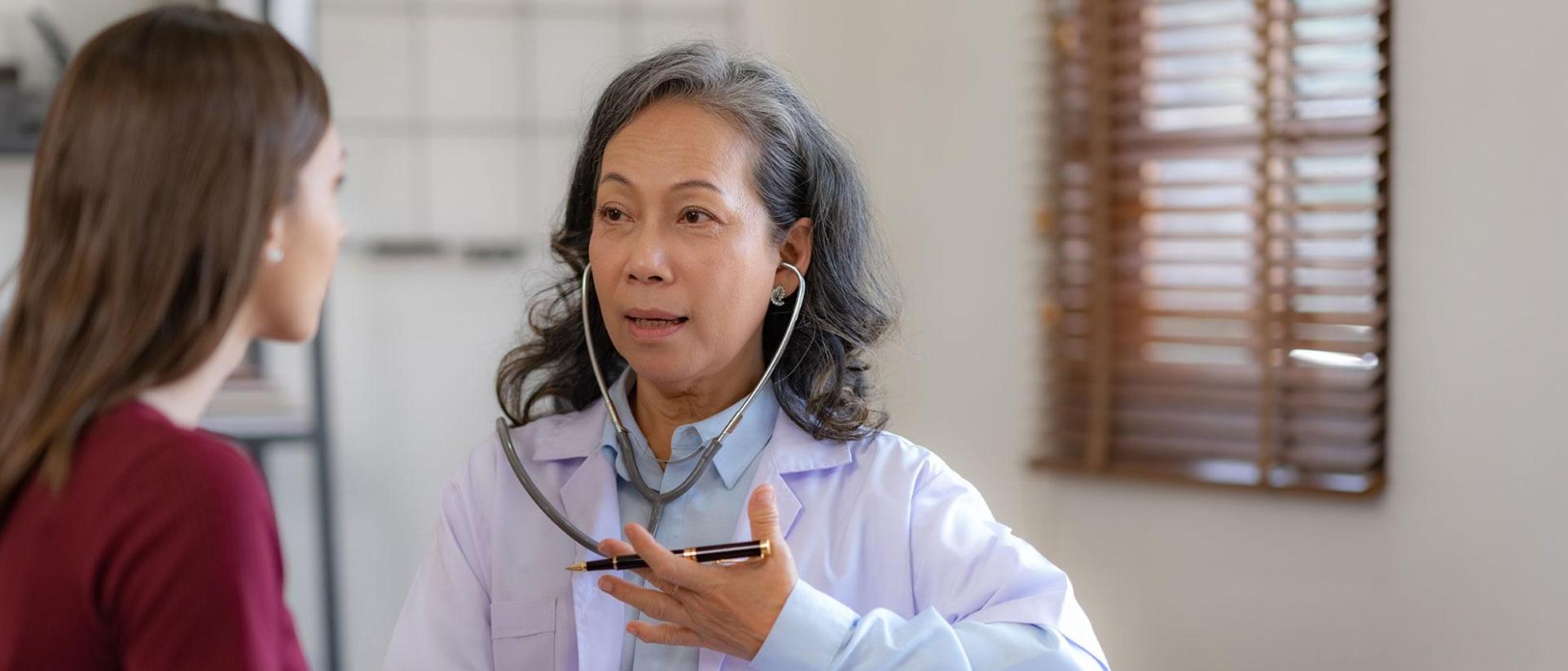 Asian nurse woman with greying shoulder length hair wearing a lab coat and stethoscope in ears with her hand over her lungs, sitting with a brunette young patient in dark red shirt.
