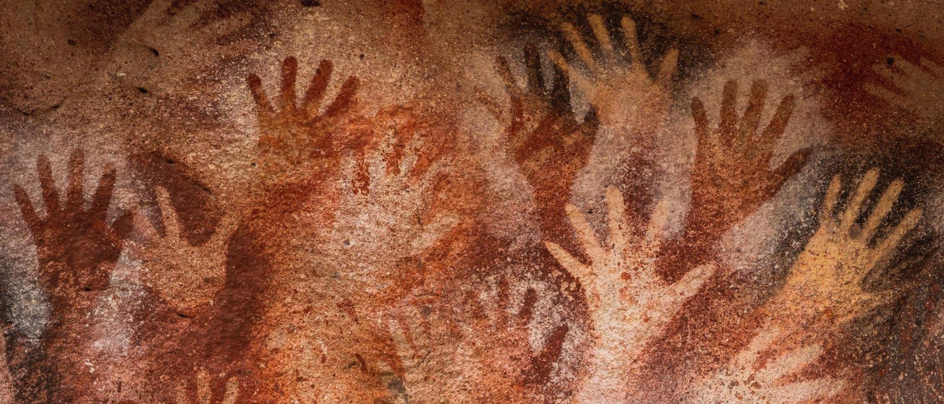 Many handprints outlined with dark orange and red paint on cave wall.