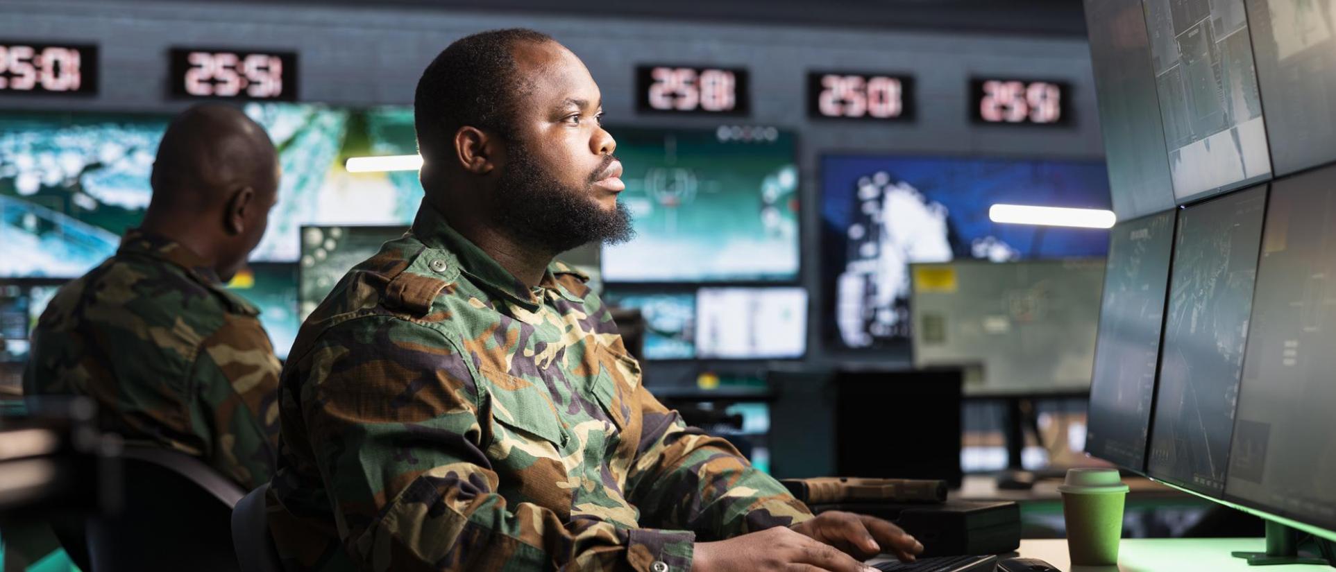Man wearing camo sits in control room looking into many computer monitors.