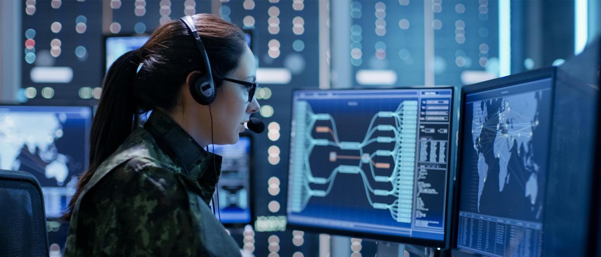 Woman wearing camo, glasses and headphones, looks into multiple computer monitors showing complex maps.