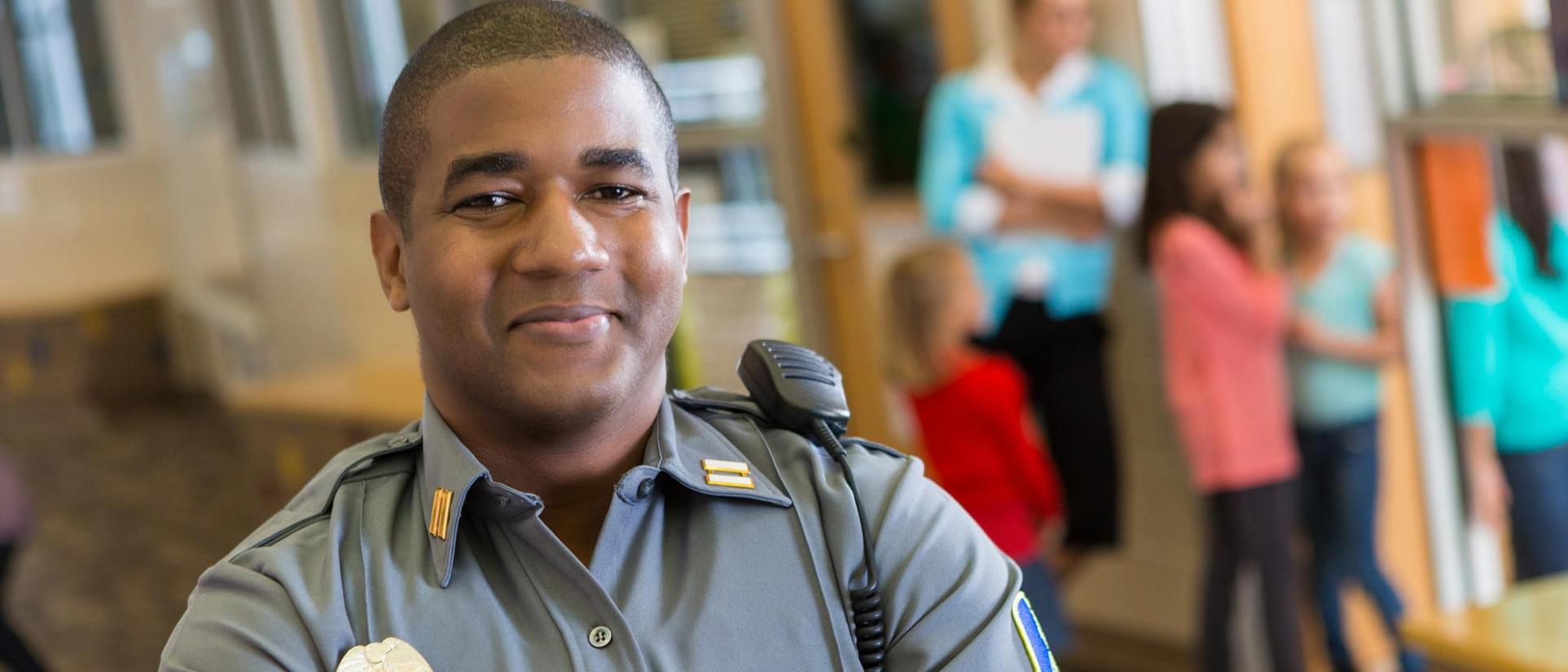 A man wearing a law enforcement uniform with a radio on his shoulder smiles at the camera while children gather in the background.