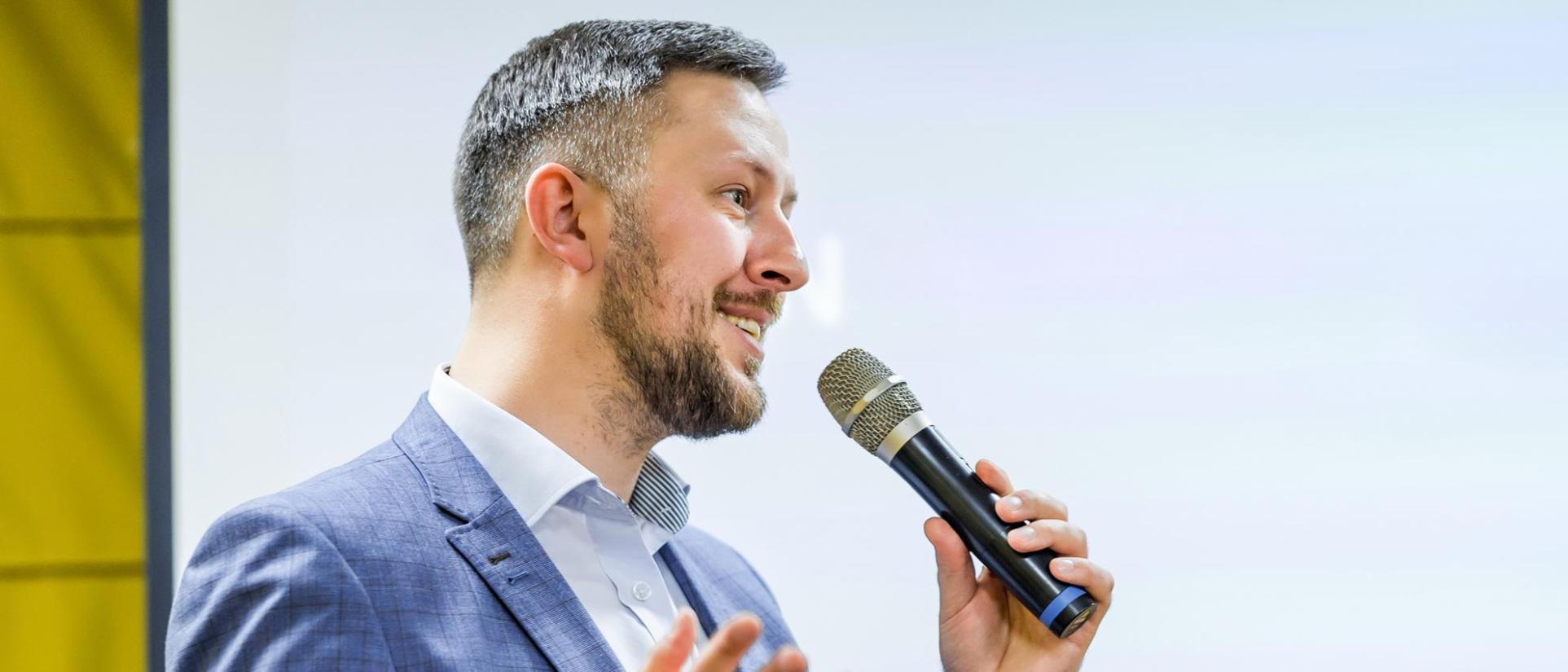 Man smiles with microphone in hand, public speaking on stage.