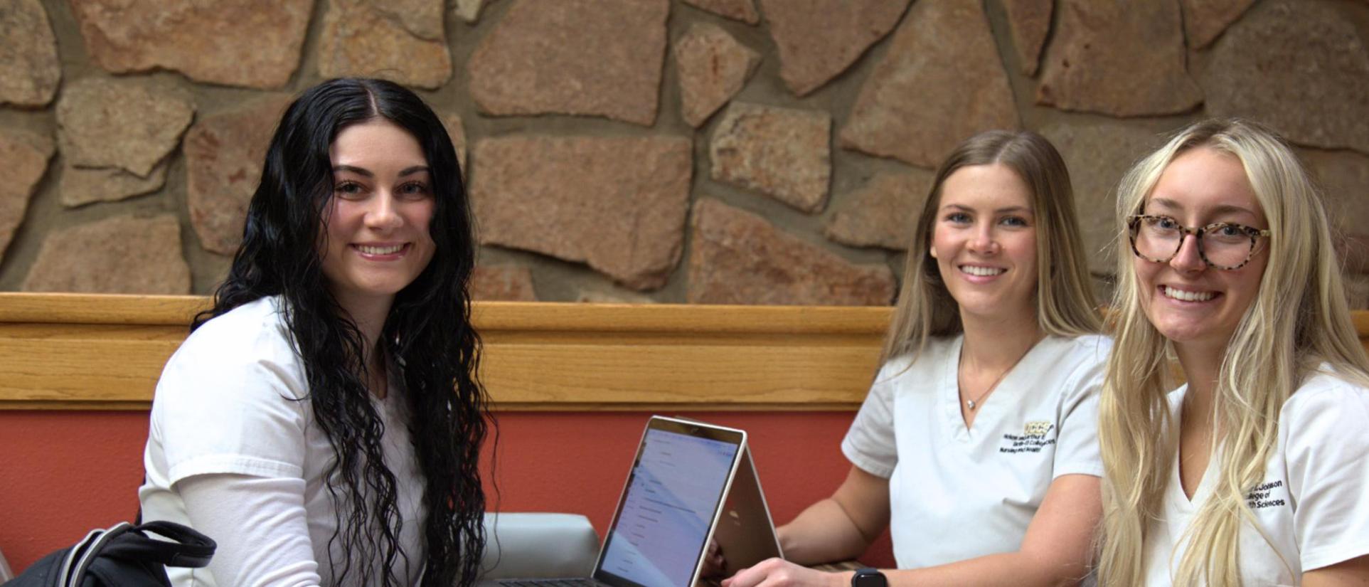 3 UCCS nursing students in white scrubs sit at table in front of laptops.