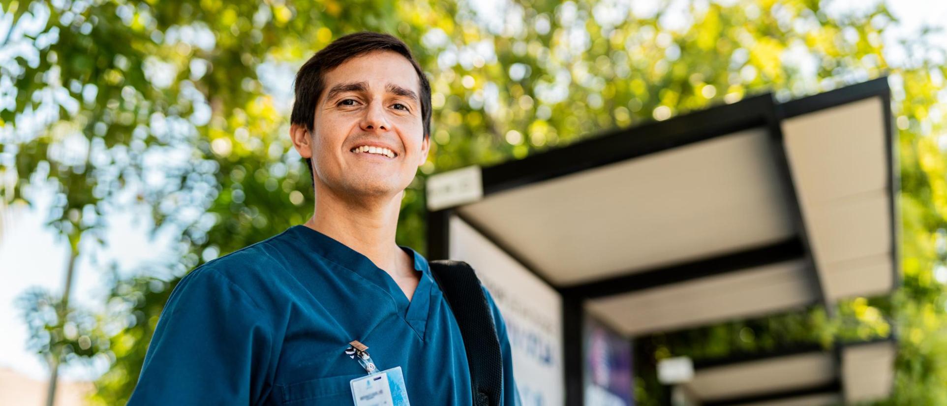 Nurse man with official id pass clipped to dark teal scrub shirt smiles in front of leafy green trees.