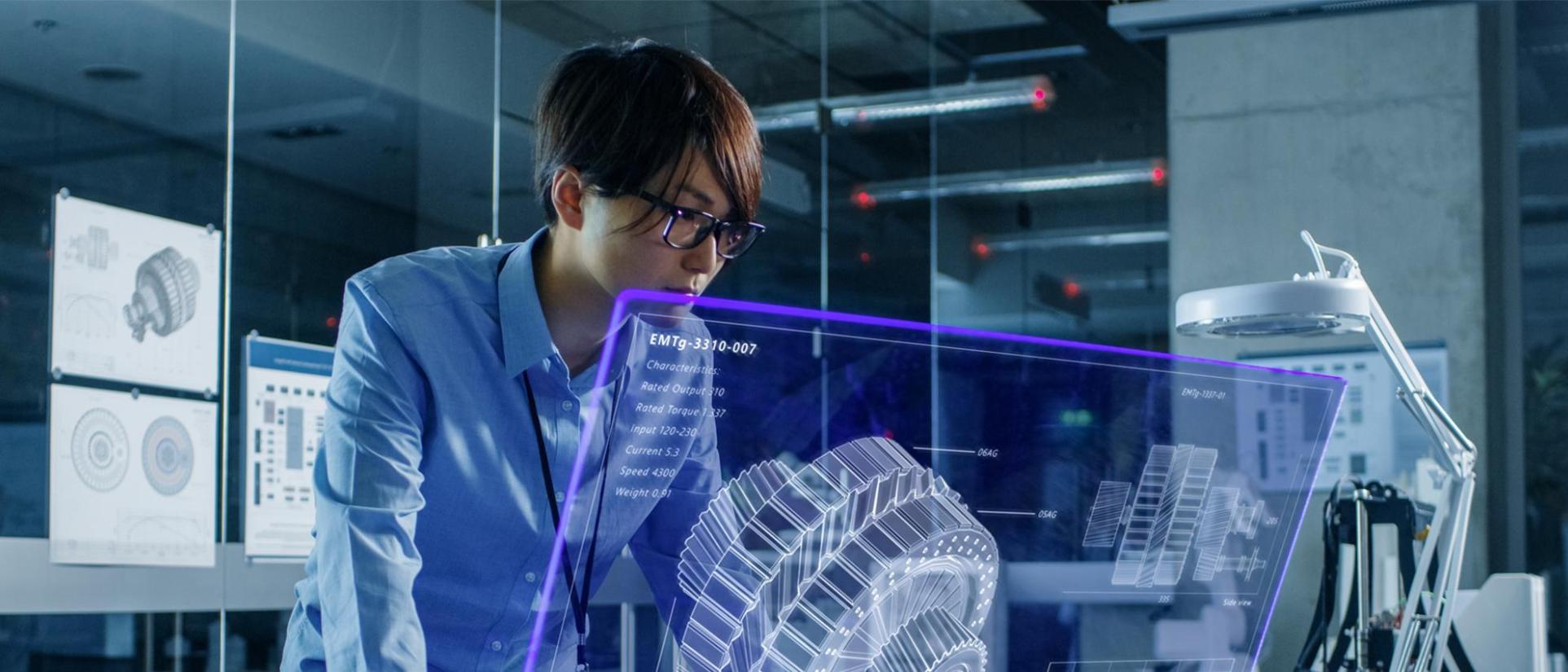 Young man with black hair and glasses, looks into a blue holographic screen with a large engineering cog system.