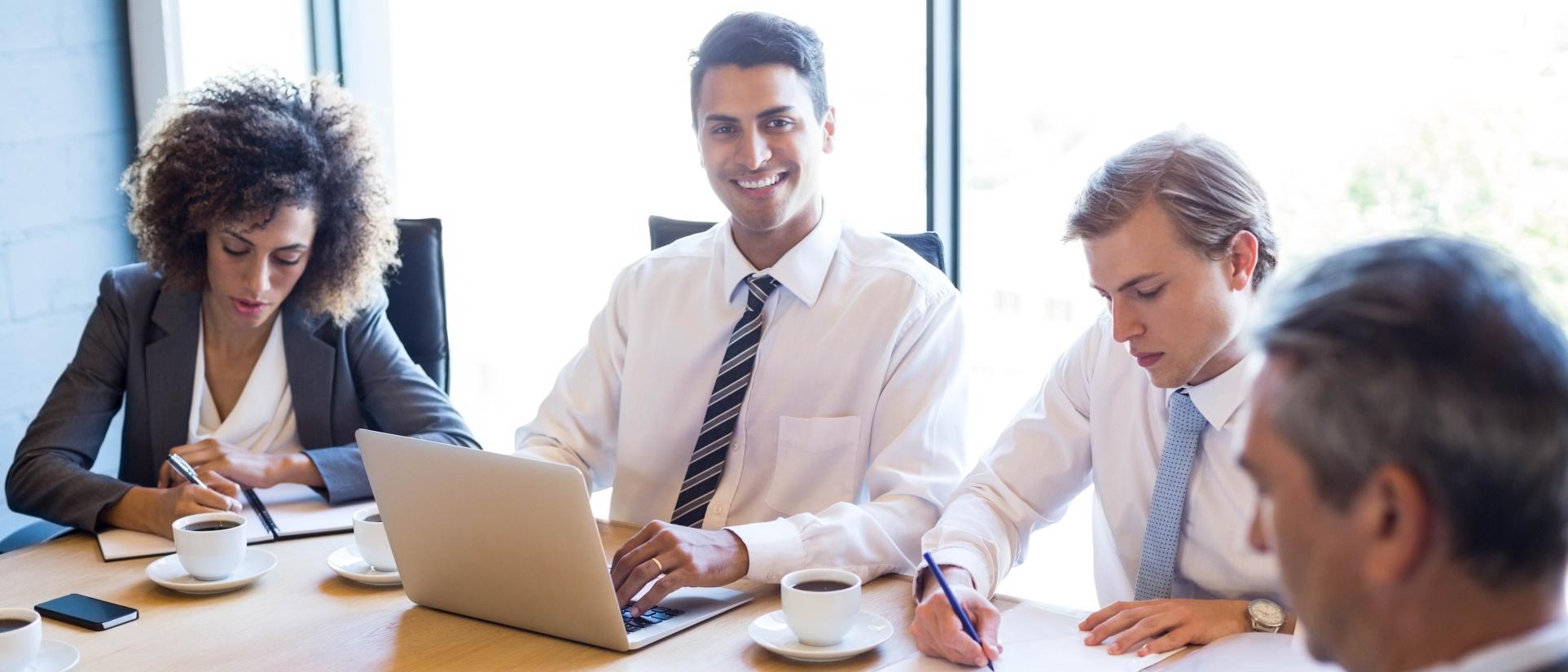 Man wearing a tie, sits at a table smiling, typing on a laptop with other well-dressed workers around him, busy writing on papers.