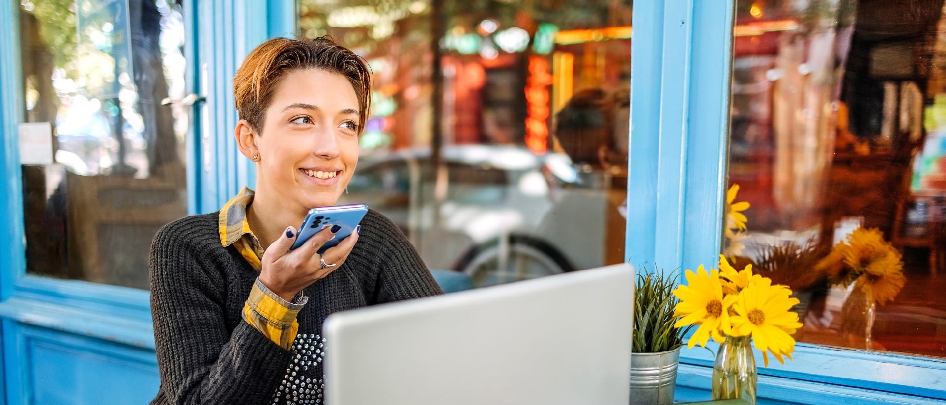 Young woman with short hair holds a cell phone up to her lips, while sitting at a table outside, in front of a laptop.