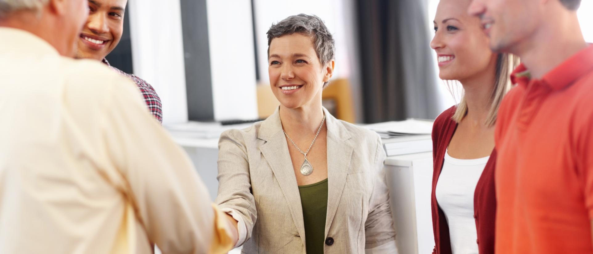 Woman wearing beige blazer and silver tear drop shaped necklace, with short greying hair, smiles as she shakes an older man's hand with other people surrounding them in an office setting.
