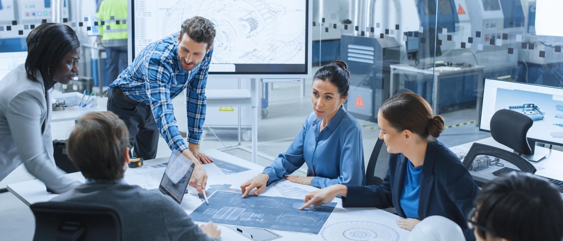 Man in plaid shirt leans over a table surrounded by a group of colleagues all looking at a large blueprint that he's pointing to.