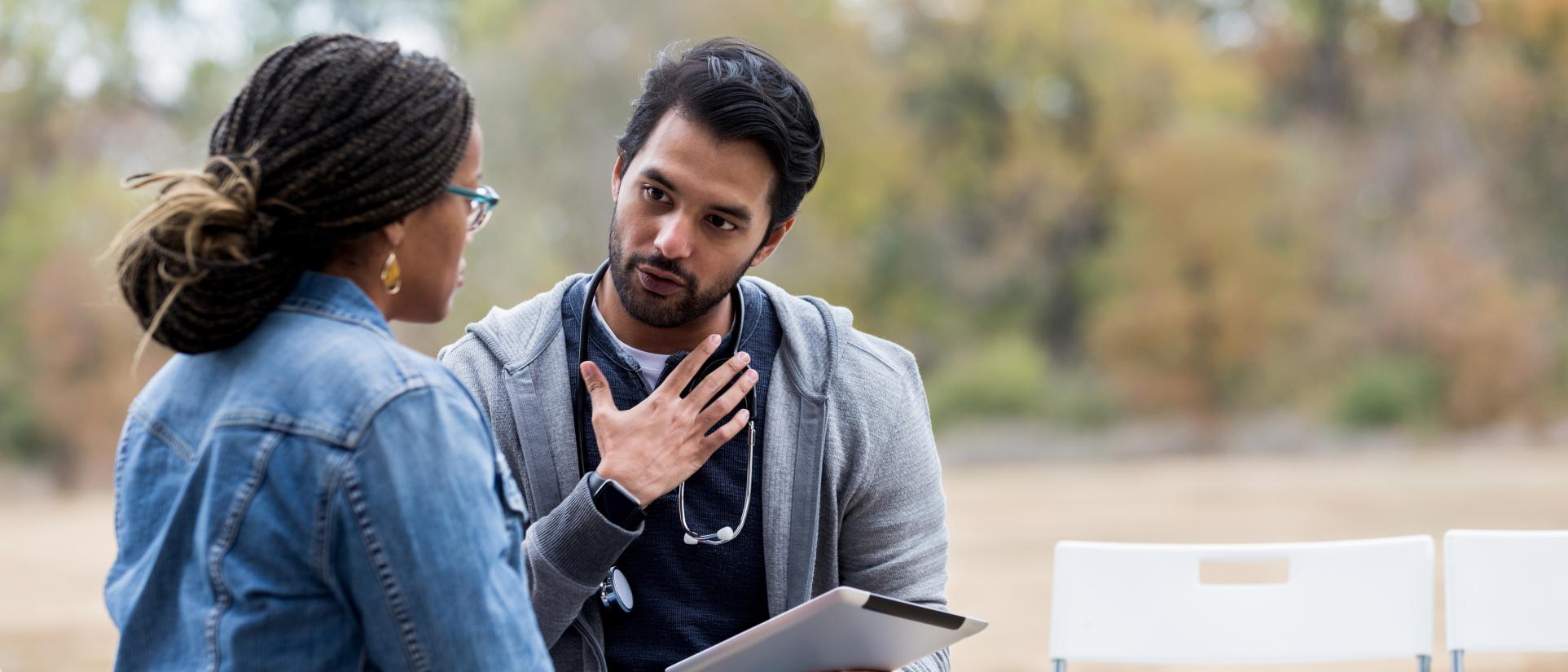 Dark haired man wearing hoodie sweatshirt and stethoscope around his neck, sits outside with woman, while gesturing to his heart and holding a tablet.