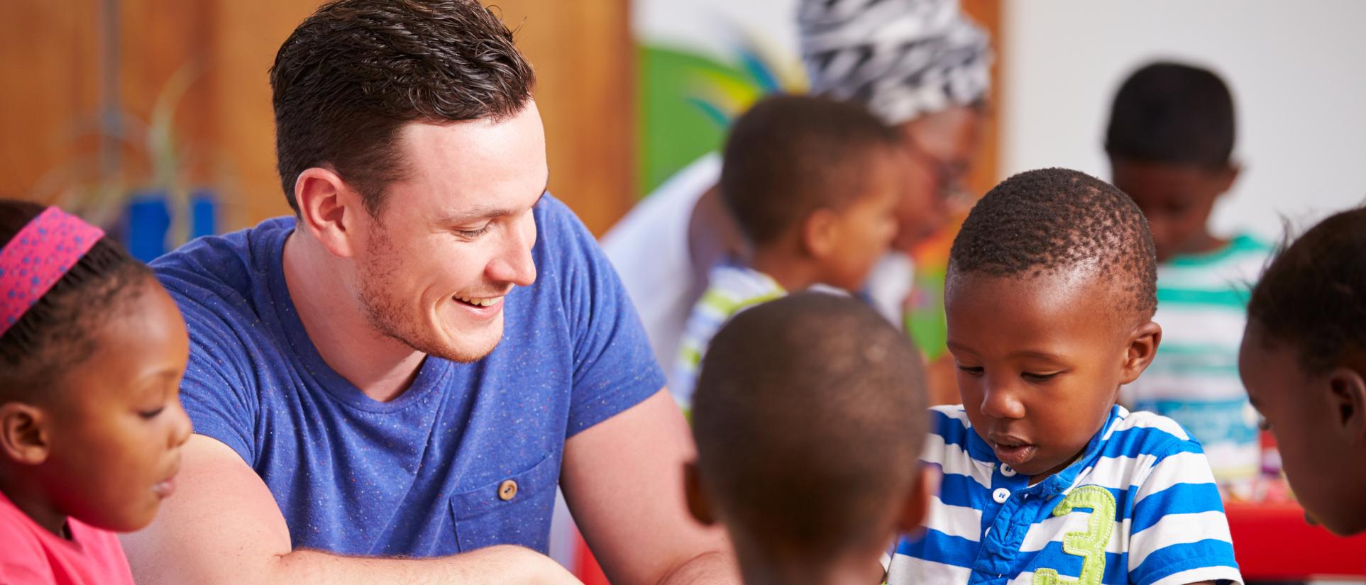Young teacher man smiles at many children around him in a classroom.