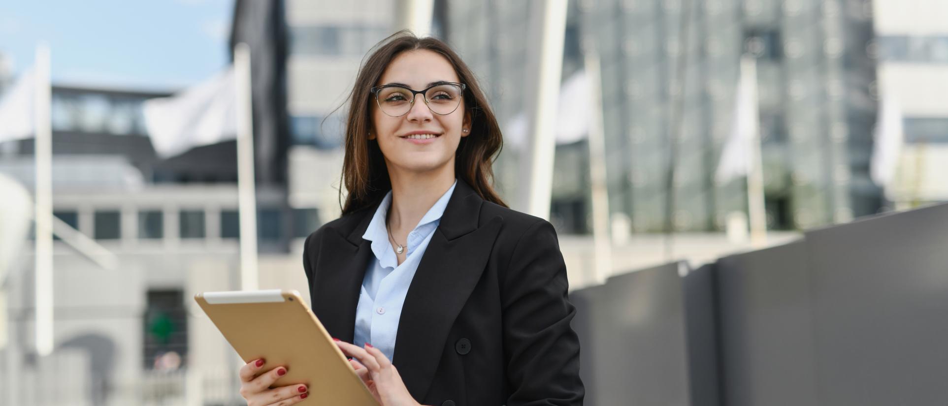 Woman smiling, wearing glasses and blazer with tablet in her hands. She stands in front of flags, with buildings in the background.