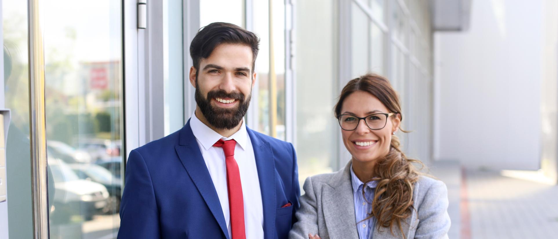 Outside of a glass-lined building, a hansom man with thick beard, wearing a blue suit with red tie, smiles next to a happy, brunette, woman next to him, wearing a grey blazer.