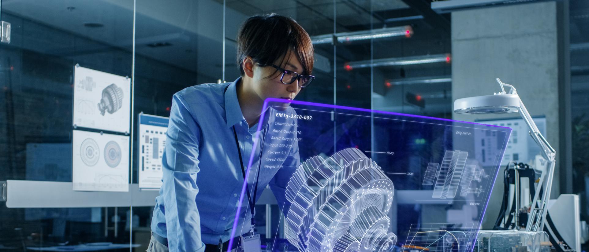 Young man with black hair and glasses, looks into a blue holographic screen with a large engineering cog system.