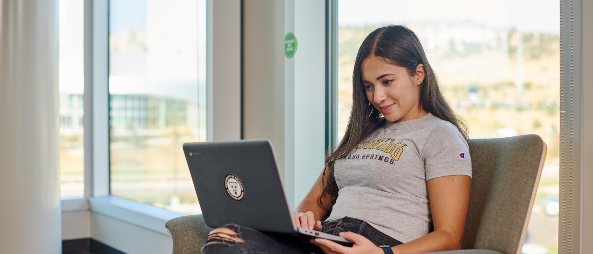 Young, female student with long, brunette hair, smiles as she looks into a laptop computer on her lap, sitting in front of large windows.