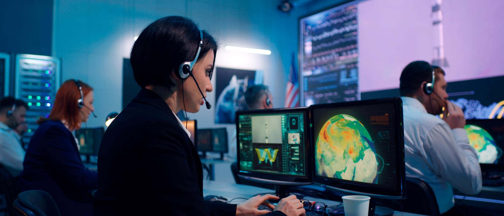 Woman in control room wearing a headset, looking into computer monitor showing planet earth with a large screen in the background showing a rocket being launched.