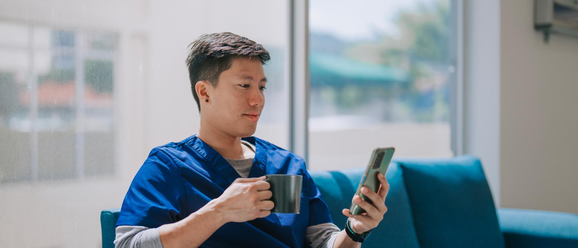 Man wearing blue scrubs, holding coffee cup, reads phone in-hand.