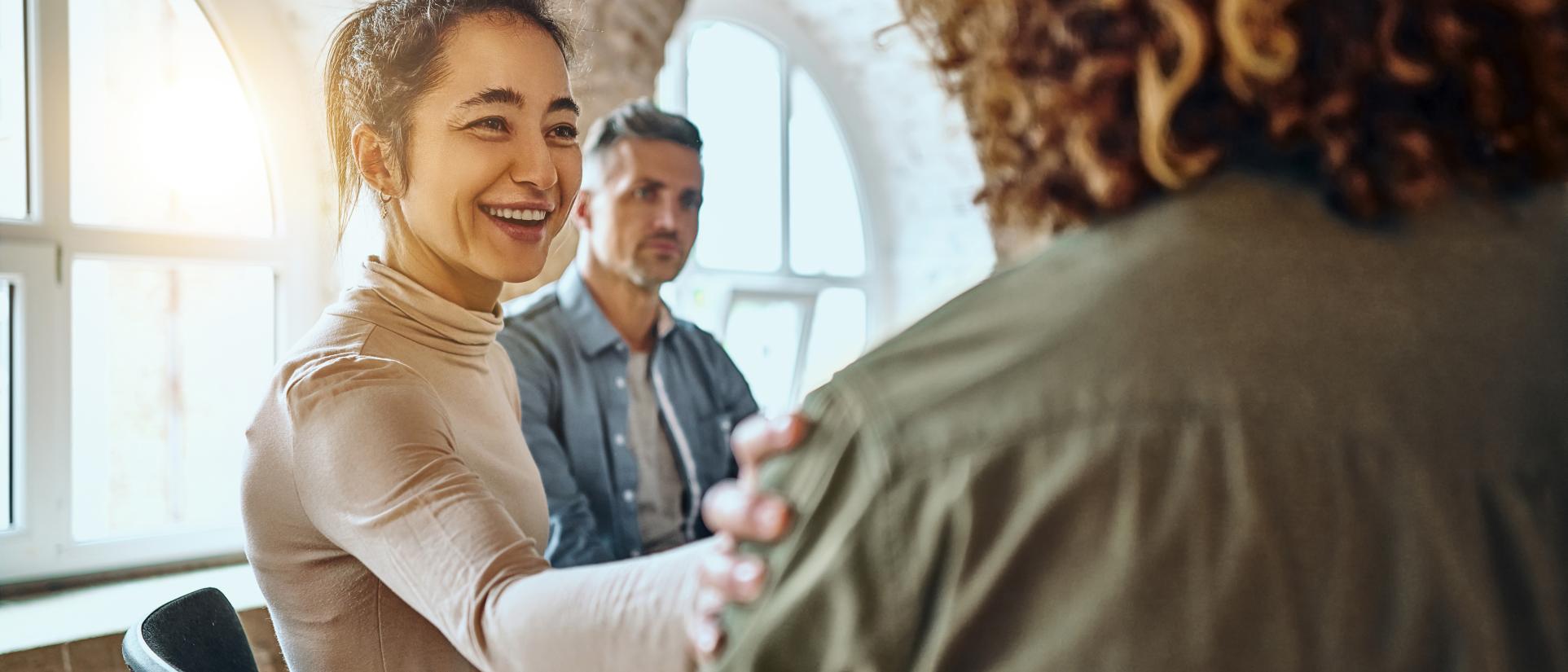 Woman with dark hair smiling and putting a helping, caring, understanding hand on a man's shoulder next to her. Other people in a round chair group gathering, with shining rays of light washing in through the window in the background.