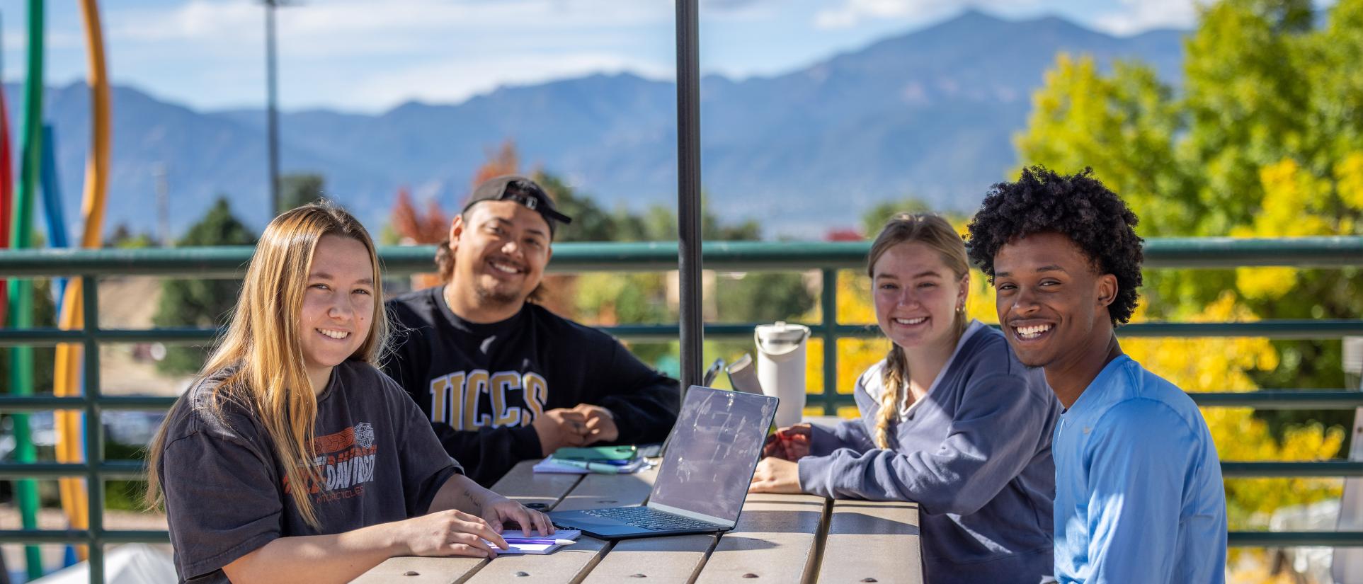 Smiling group of students sit at outdoor table with laptop. Green trees and beautiful blue mountain range in the background.