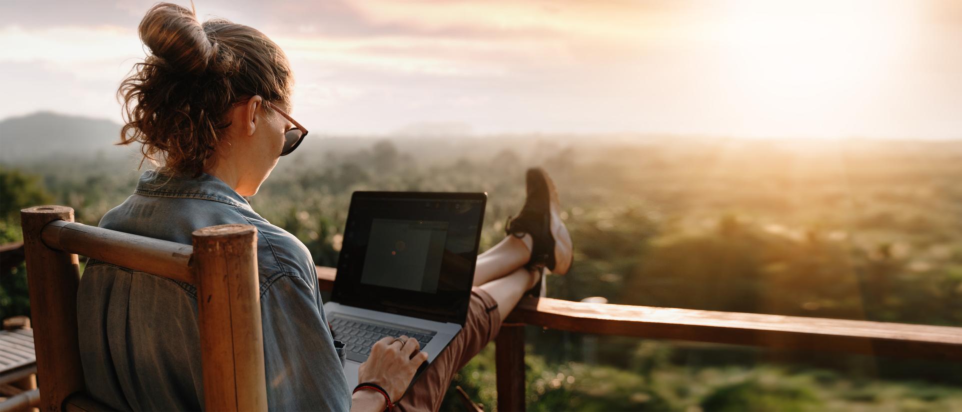 Woman sits in wooden chair outside with feet up and crossed on the railing of a deck overlooking a beautiful scenic vista with sun setting in the distance. She's wearing a denim jacket and sunglasses, looking into laptop.