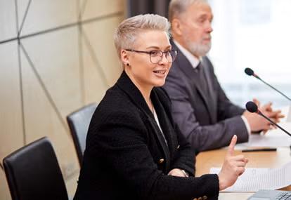 Woman in black blazer with short light hair with her finger up sits at table next to older man, both with microphones 