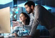 Woman sitting in front of large silver computer monitor with a man behind her pointing at the screen which is projecting holographic images of earth continents with map grid.