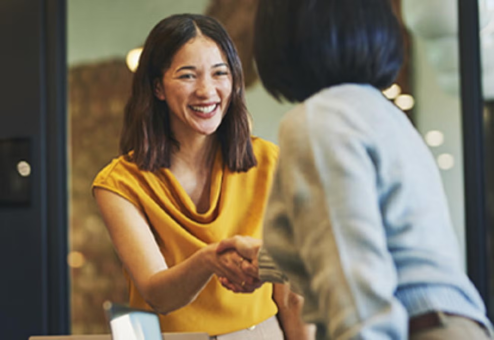 Smiling young woman in gold shirt shakes employer woman's hand.