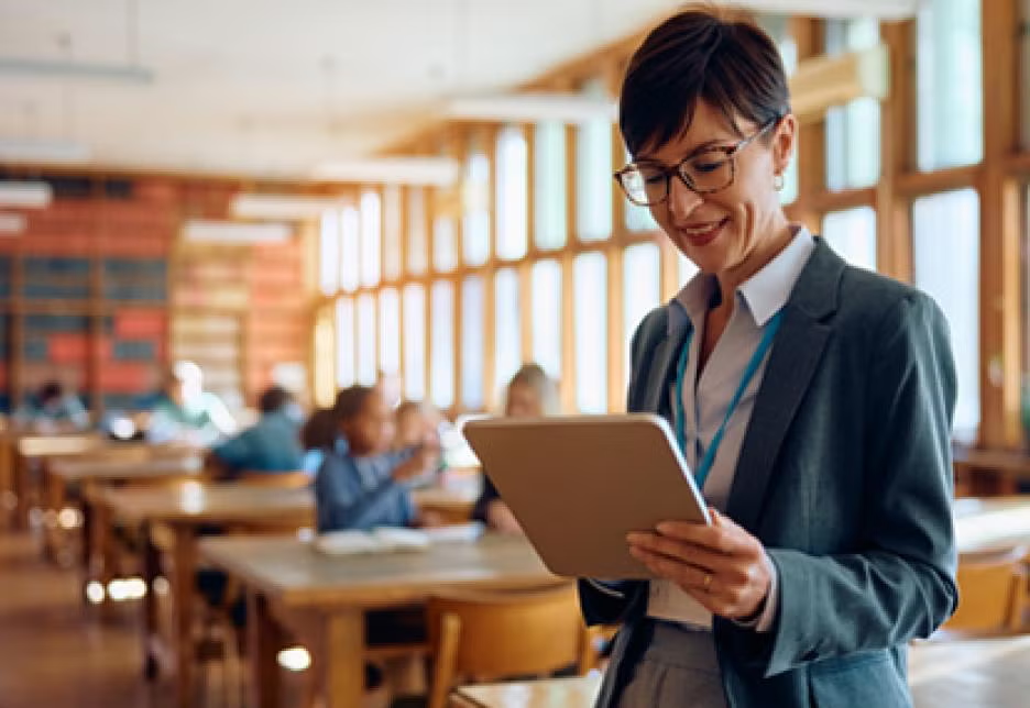 Teacher woman looks down into tablet standing in front of a classroom of kids at their desks.