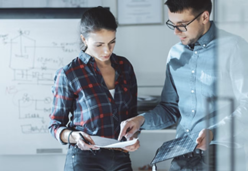 Man and woman stand if front of white board pointing at tablet.