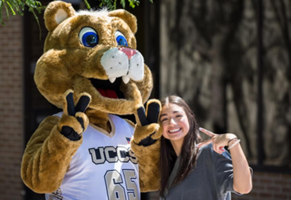 Smiling female college student with the UCCS mascot Clyde the Mountain Lion, both holding up 2-finger peace signs.