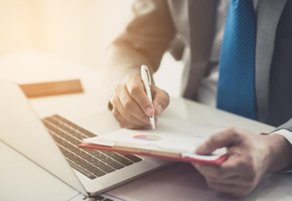 Man's hands holding a pen and paperwork showing a pie chart with laptop in the background.