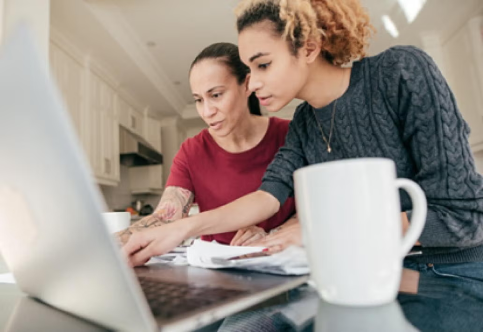 Mother an daughter look into laptop with paperwork and coffee cup on their counter.