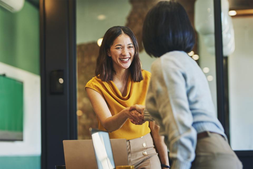 Smiling, dark-haired woman in golden yellow blouse shakes another woman's hand, over laptop computers.