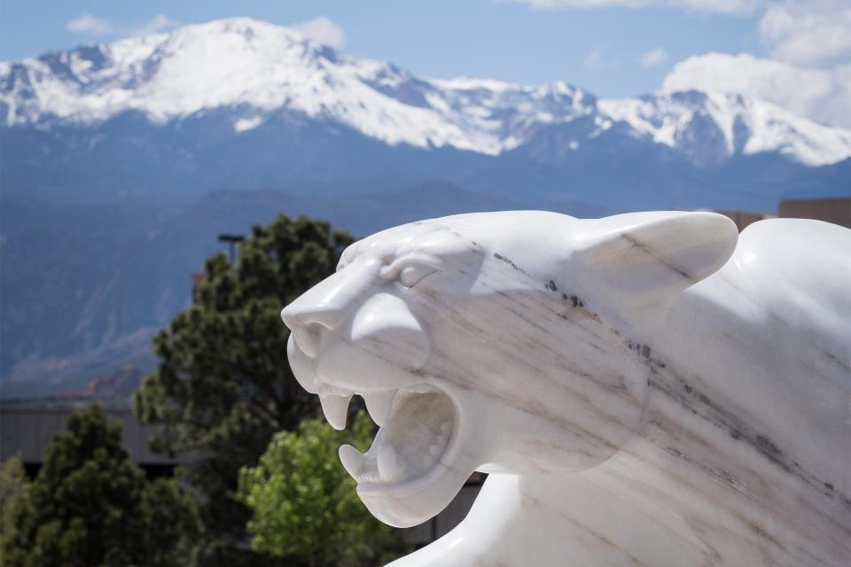 Beautiful snow-capped mountain range in the background, marble statue of a mountain lion in the foreground, evergreen trees in between.