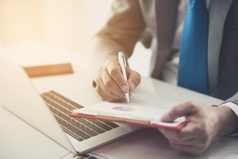 Laptop computer with man's hands holding clipboard and pen, wearing blue tie.