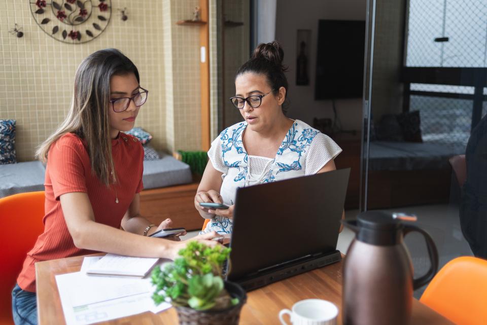 Dark haired mother and daughter, both wearing glasses, sitting at a laptop with paperwork on the table and mother typing at calculator.