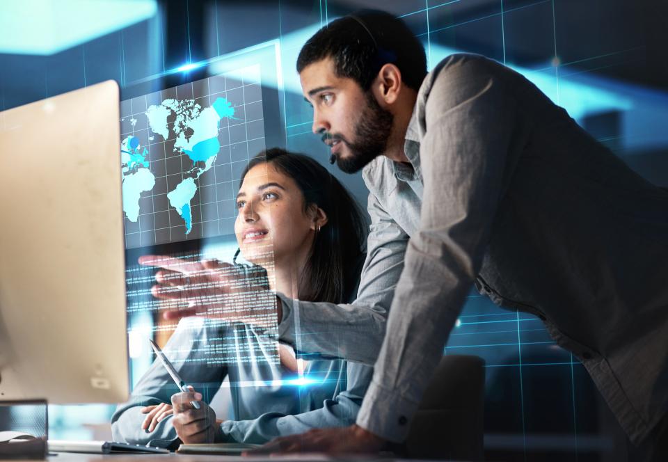 Woman sitting in front of computer looking at holographic map. Man with beard bends over her shoulder pointing at the screen.