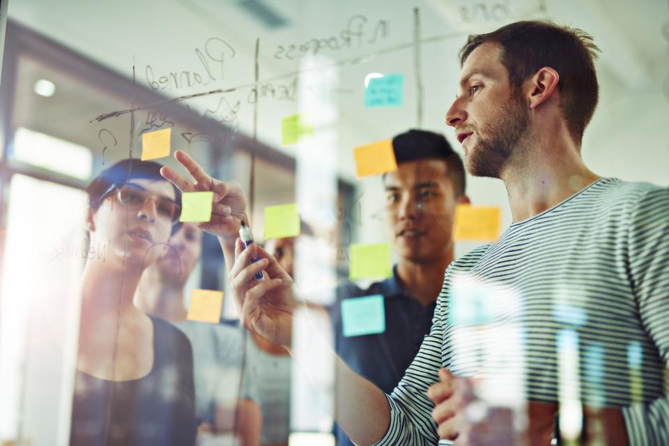Man surrounded by group of diverse co-workers behind glass. He's drawing with erasable marker on the glass around lots of neon colorful sticky notes.